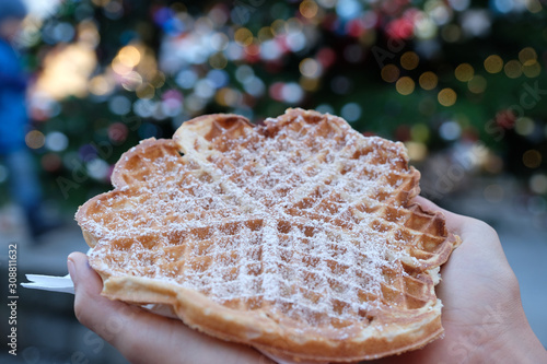 Selective focus two hands holding one tasty waffle with icing (powdered )sugar. Blurred colourful Christmas balls and multi-colored bokeh background (at German Christmas market or Weihnachtsmarkt).