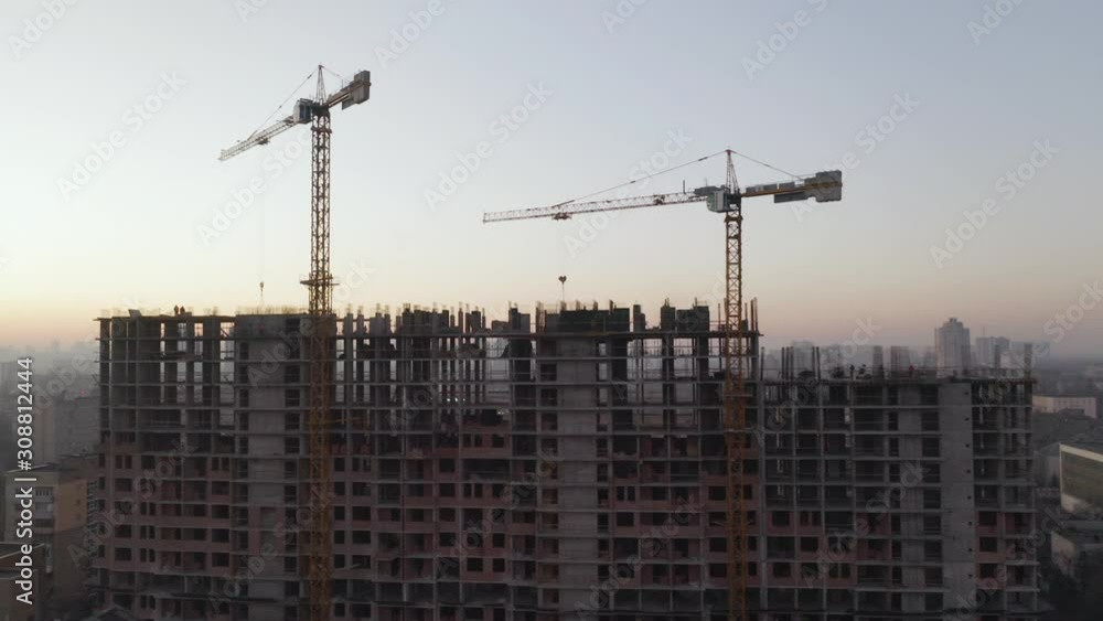 In the Background Working Crane and city. Aerial Shot of the Building in the Process of Construction. Labor working in big construction site In the Background Working Crane and city. Aerial Shot of the Building in the Process of Construction. Labor working in big construction site