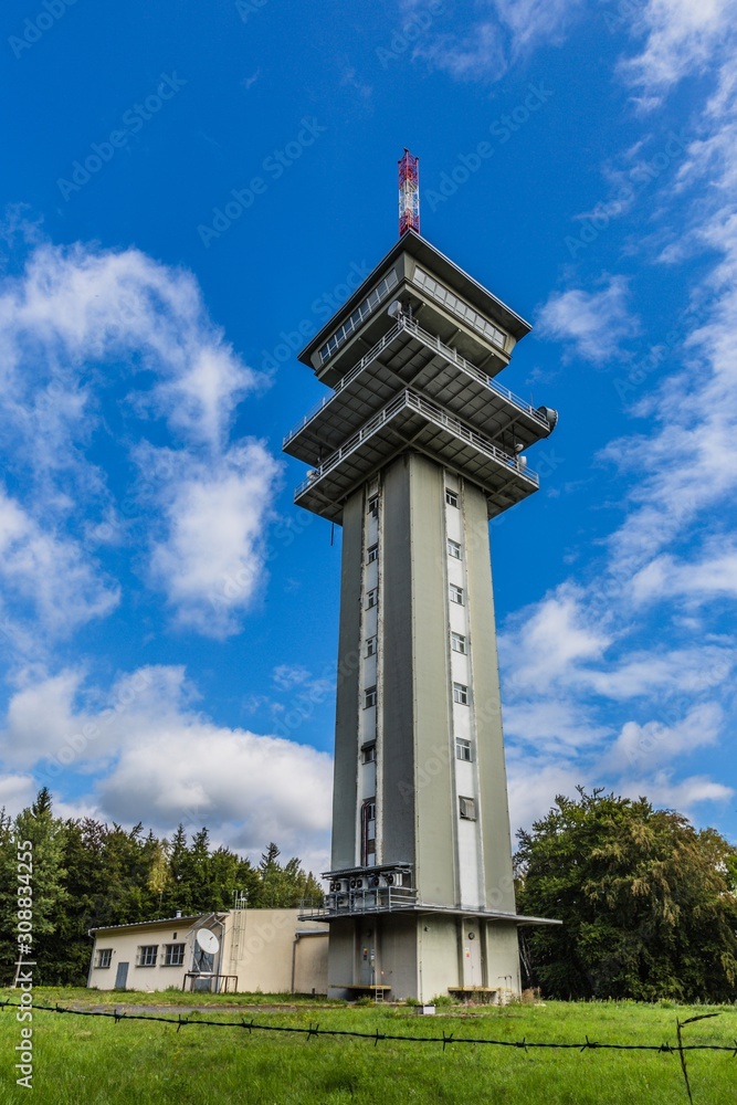 Zelena hora, Pelhrimov / Czech Republic - September 13 2019: Vertical image of television communication tower, a tall grey building on a sunny day with blue sky and clouds. Green trees and grass.