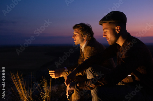 Two men are talking sitting near flaming campfire in dusk time. Gentlemen has sincere conversation, share memories and drink whiskey. Sunset sky on background