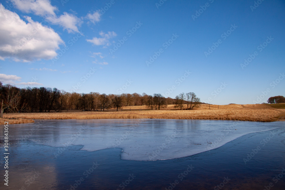 Beautiful landscape with a lake almost covered with the first thin ice. Winter or spring scene on a sunny day with clear blue sky, white clouds and a forest in the distance. Image. Beautiful landscape with a lake almost covered with the first thin ice. Winter or spring scene on a sunny day with clear blue sky, white clouds and a forest in the distance. Image.