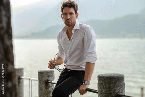 Stylish man wearing white shirt, holding sunglasses and looking at camera. Posing over lake view