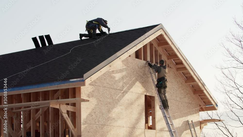 Residential house under construction. One worker is covering the wooden frame with plywood using hydraulic hammer, another one is making ridge on the roof in safety harness Residential house under construction. One worker is covering the wooden frame with plywood using hydraulic hammer, another one is making ridge on the roof in safety harness