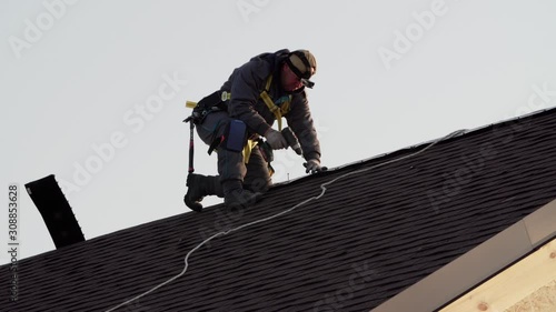 Professional tiler in safety harness is making roof ridge with hydraulic hammer. Works at height. Wooden frame house under construction
