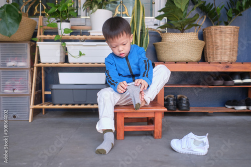 Cute little Asian 3 years old toddler kindergarten kid sitting near shoe rack near front door of his house and concentrate on putting on his own socks, Encourage Self-Help Skills in Children concept