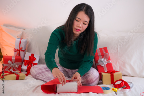 Christmas holiday lifestyle portrait of young happy and sweet Asian Japanese woman sitting on bed preparing xmas presents wrapping gift boxes and adding ribbons