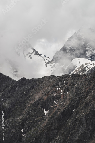 Atmospheric minimalist alpine landscape with big rocky mountain wall and snowbound peak in low clouds. Huge mountains with massive glacier. Craggy wall with snow. Majestic scenery on high altitude. © Daniil