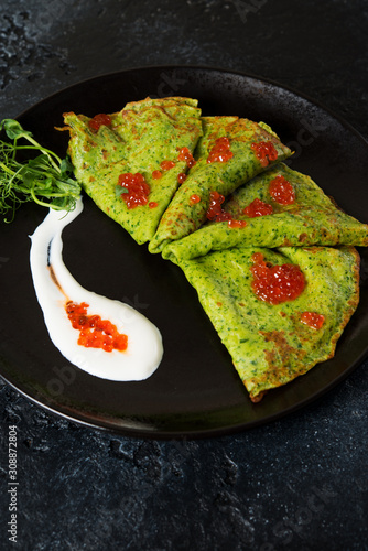 Classic thin green spinach pancakes with sour cream and red caviar in a black plate.