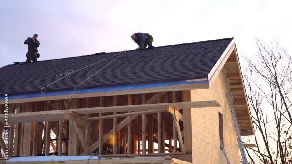 View from below of two builders in safety harness working on the roof. Wooden frame house building View from below of two builders in safety harness working on the roof. Wooden frame house building