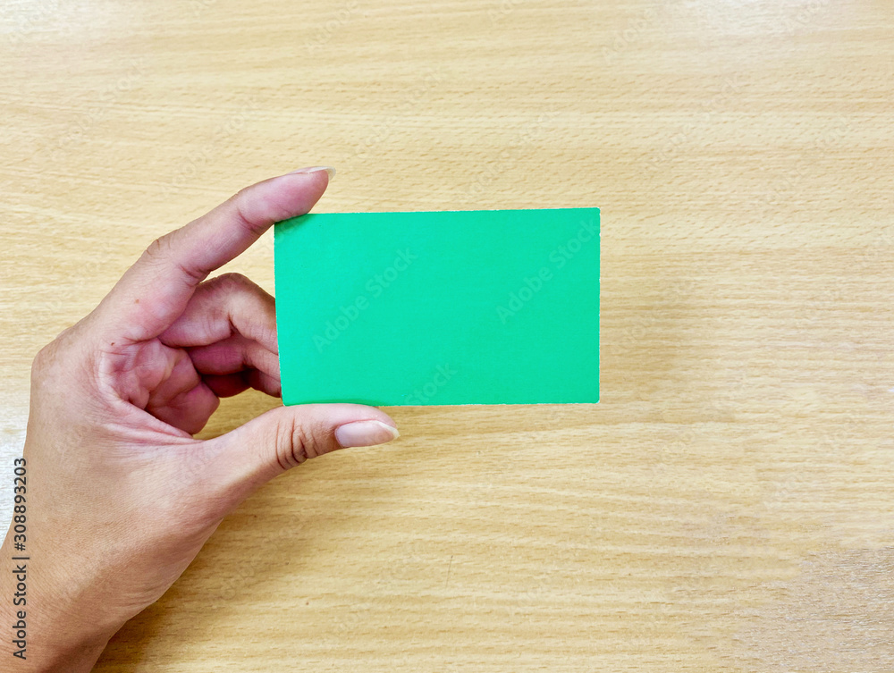The hand of an Asian woman holding a blank green business or name card and the background is light brown wood. For use in insert text, message, comment, reply or chat in advertising public relations. 