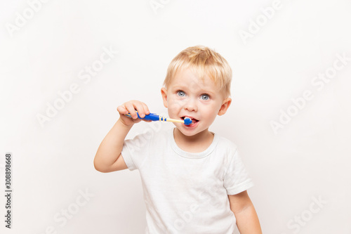little cute caucasian blond blue-eyed boy in a white t-shirt brushes his teeth on a white background with copy space