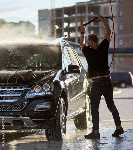 Handsome man holding a hose and washing his black car with a water jet, water drops and side of the car are shining on the setting sun