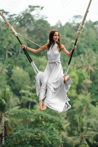 Tanned beautiful woman in a long white dress with a train, riding on a swing. In the background, a rainforest and palm trees. Vertical orientation