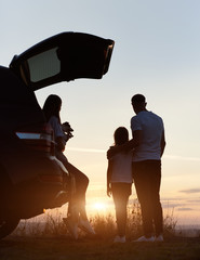 Silhouette of the family of three enjoying the sunset on the top of hill outside the city, woman is sitting in the car trunk, holding jack russell terrier, man is standing embracing his daughter