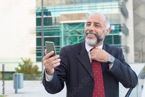 Happy positive mature business leader adjusting tie and using smartphone. Elderly man in formal suit and tie standing outside in city. Mobile phone using concept