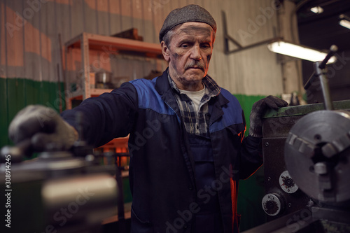 Senior serious man in work wear standing and looking at the work of machine in the factory