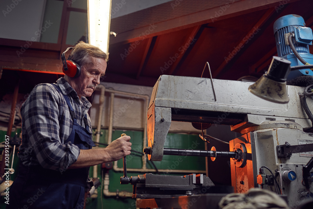 Serious senior operator wearing headphones standing at the lathe and operating it in the factory Serious senior operator wearing headphones standing at the lathe and operating it in the factory