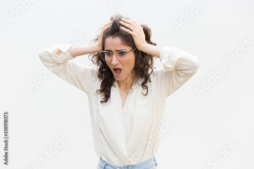 Shocked upset woman in glasses gasping, holding head and staring away at copy space. Wavy haired young woman in casual shirt standing isolated over white background. Failure concept