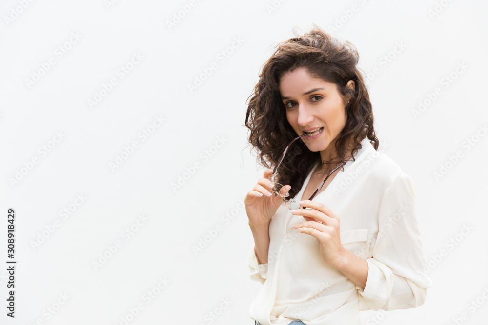 Positive playful student girl biting glasses temple. Wavy haired young woman in casual shirt standing isolated over white background. Attractive woman portrait concept Positive playful student girl biting glasses temple. Wavy haired young woman in casual shirt standing isolated over white background. Attractive woman portrait concept