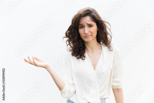 Doubtful woman pointing hand at empty copy space and shrugging. Wavy haired young woman in casual shirt standing isolated over white background. Advertising concept