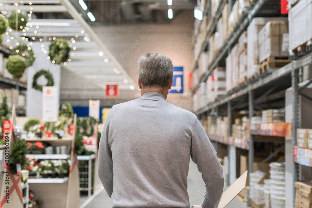 Tall gray-haired man shopper walking with trolley in a large hardware store before christmas. Back view Tall gray-haired man shopper walking with trolley in a large hardware store before christmas. Back view