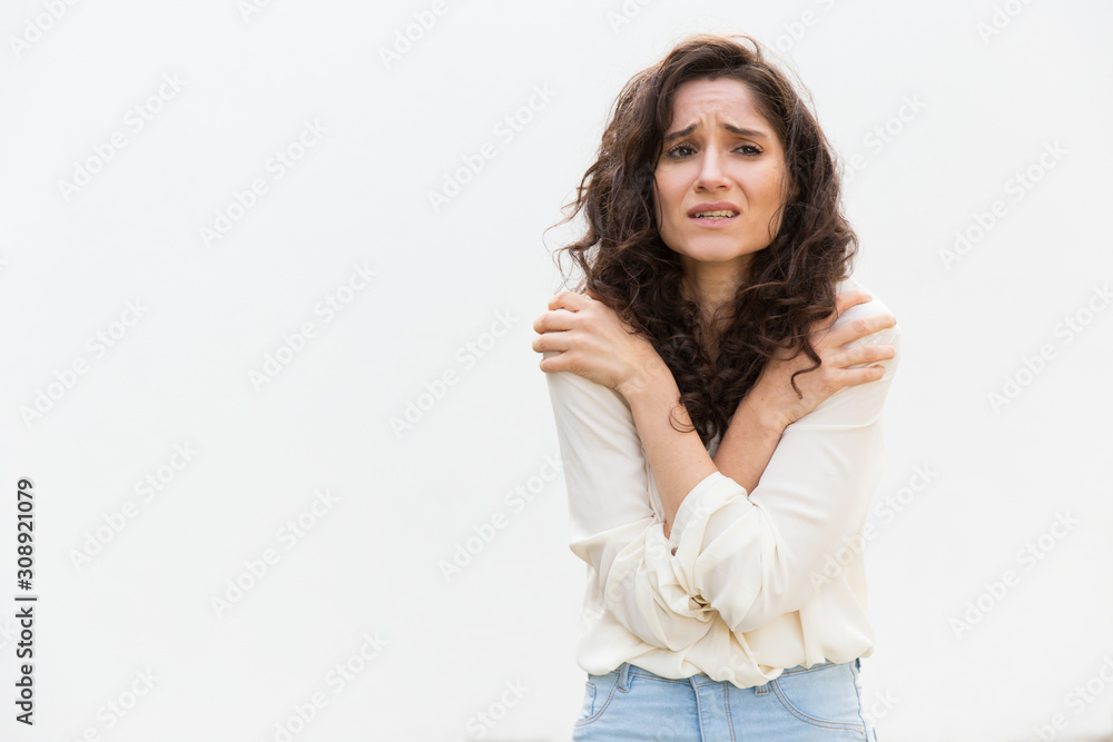 Desperate upset woman freezing, trembling, hugging herself for warm. Wavy haired young woman in casual shirt standing isolated over white background. Fever or fear concept Desperate upset woman freezing, trembling, hugging herself for warm. Wavy haired young woman in casual shirt standing isolated over white background. Fever or fear concept
