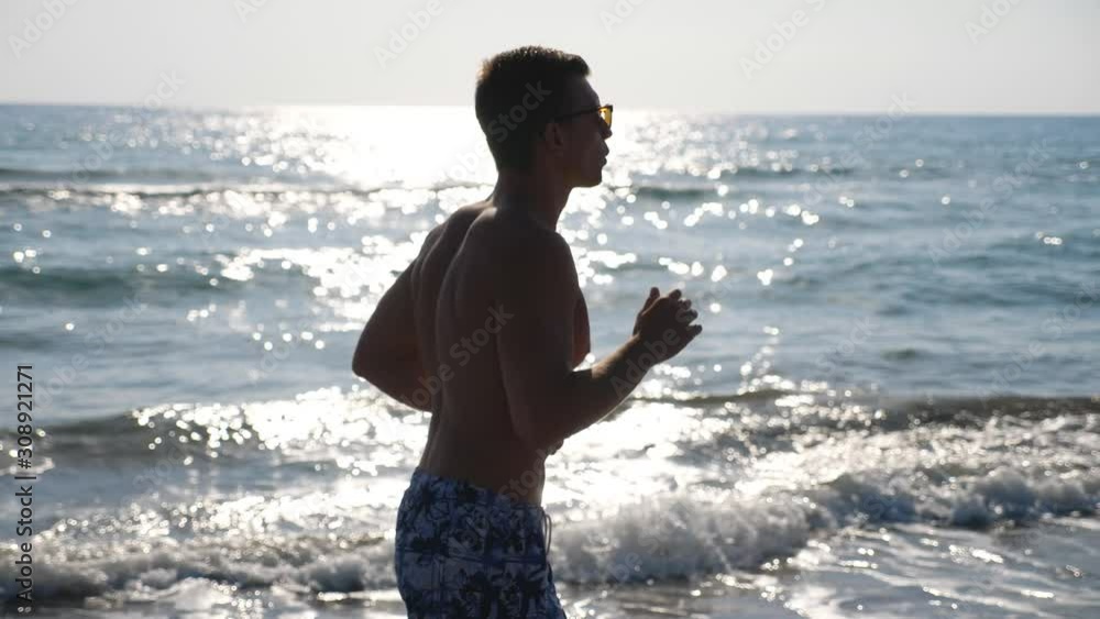 Athletic handsome guy jogging along sea coast at morning. Young sporty man running through ocean beach. Close up of male sportsman doing sport on seashore. Healthy active lifestyle. Slow mo Side view Athletic handsome guy jogging along sea coast at morning. Young sporty man running through ocean beach. Close up of male sportsman doing sport on seashore. Healthy active lifestyle. Slow mo Side view