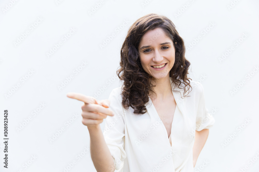 Joyful positive woman pointing finger away at copy space. Wavy haired young woman in casual shirt standing isolated over white background. Advertising concept Joyful positive woman pointing finger away at copy space. Wavy haired young woman in casual shirt standing isolated over white background. Advertising concept