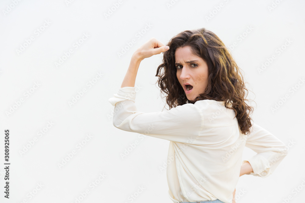 Side of severe strong feminist woman flexing bicep, showing hand muscle. Wavy haired young woman in casual shirt standing isolated over white background. Girl power concept Side of severe strong feminist woman flexing bicep, showing hand muscle. Wavy haired young woman in casual shirt standing isolated over white background. Girl power concept