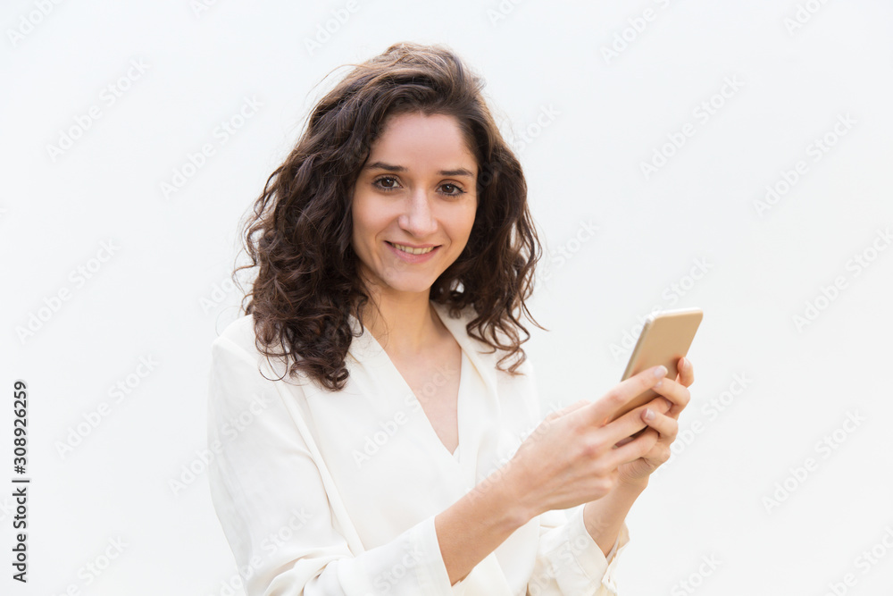 Cheerful positive female smartphone user holding device, looking at camera, smiling. Wavy haired young woman in casual shirt standing isolated over white background. Phone user concept