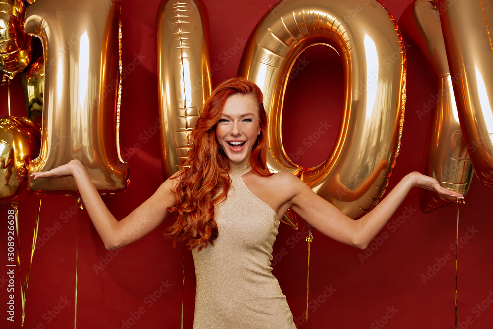 Joyful happy european woman with red haired wearing gold dress posing with wonderful smile over red background.