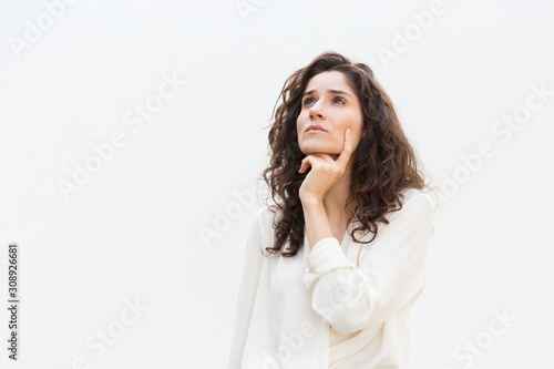 Pensive female customer leaning chin on hand and looking up at copy space. Wavy haired young woman in casual shirt standing isolated over white background. Special offer concept