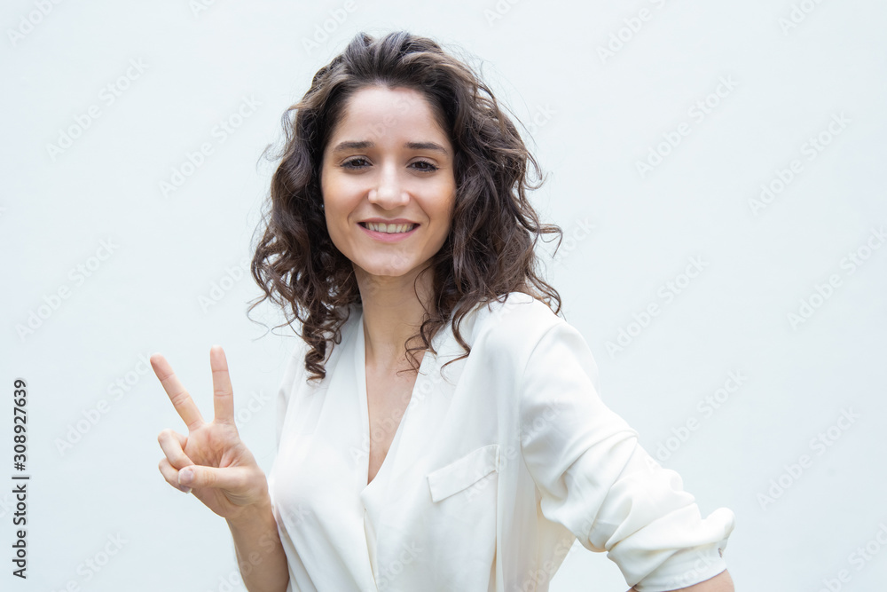 Happy carefree woman making peace sign with two fingers. Wavy haired young woman in casual shirt standing isolated over white background. Victory gesture concept Happy carefree woman making peace sign with two fingers. Wavy haired young woman in casual shirt standing isolated over white background. Victory gesture concept