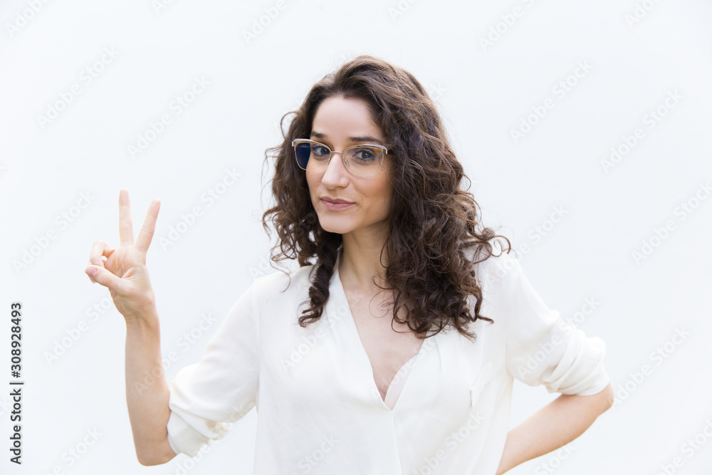 Positive woman in glasses showing victory gesture with two fingers. Wavy haired young woman in casual shirt standing isolated over white background. Peace sign concept Positive woman in glasses showing victory gesture with two fingers. Wavy haired young woman in casual shirt standing isolated over white background. Peace sign concept