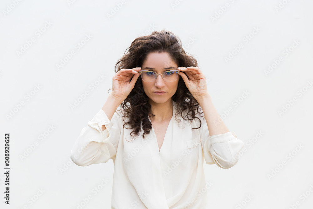Focused confident customer trying glasses on. Wavy haired young woman in casual shirt standing isolated over white background. Eyewear concept Focused confident customer trying glasses on. Wavy haired young woman in casual shirt standing isolated over white background. Eyewear concept