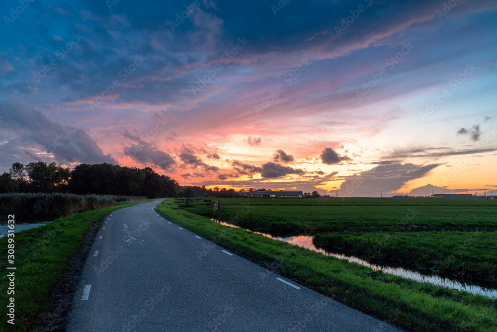 Road to sunset in Hazerswoude Rijndijk, the Netherlands. Country road during colourful sunset. Reflection in the small canals. Dutch meadows.