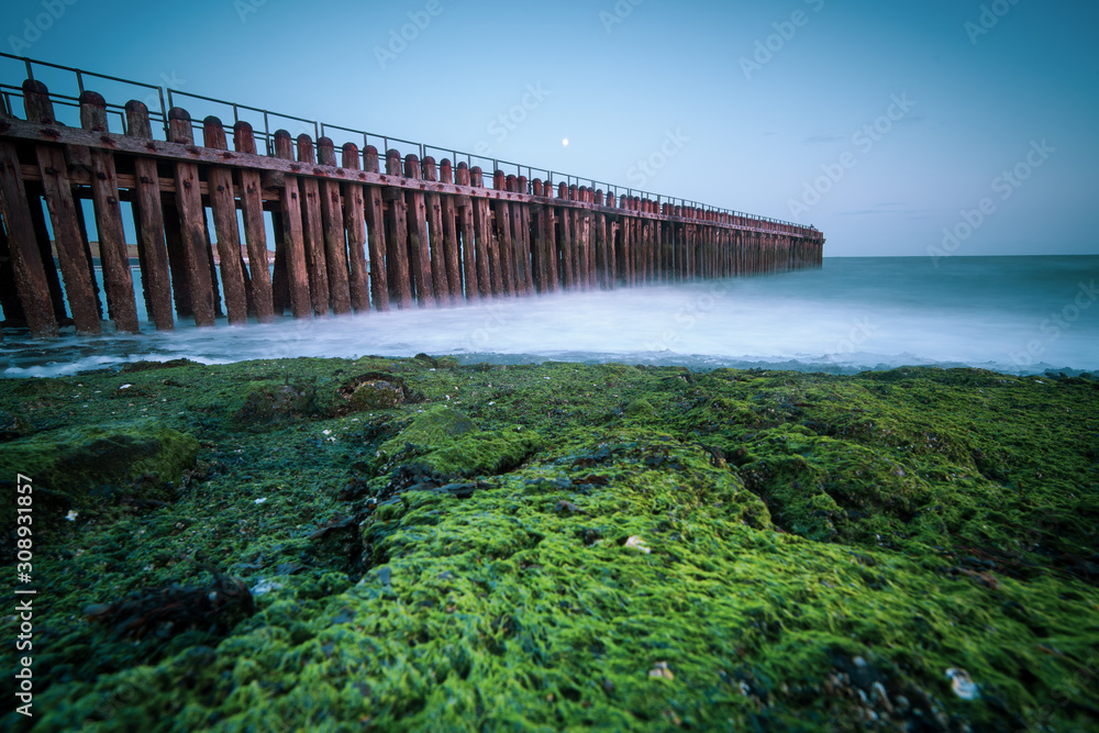 Long exposure photo during twilight with misty sea water, pier and green seaweed on the rocks
