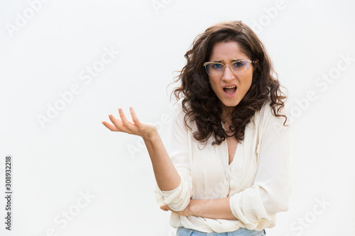Careless puzzled woman in glasses shrugging, gesturing and speaking at camera. Wavy haired young woman in casual shirt standing isolated over white background. Puzzlement concept