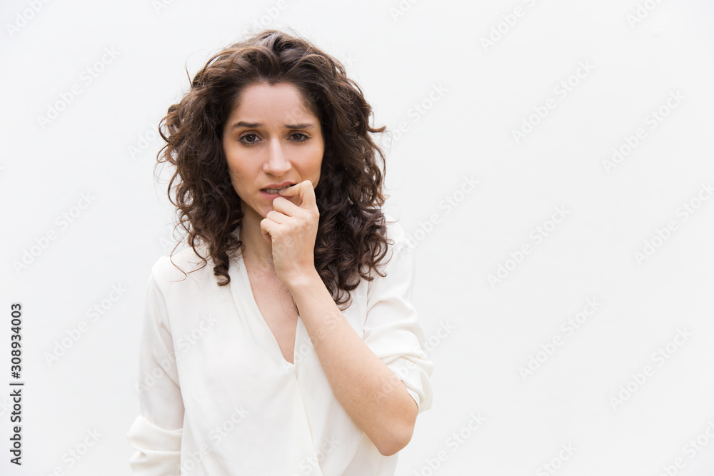 Pensive woman thinking and biting fingers and nails. Wavy haired young woman in casual shirt standing isolated over white background. Decision making concept Pensive woman thinking and biting fingers and nails. Wavy haired young woman in casual shirt standing isolated over white background. Decision making concept