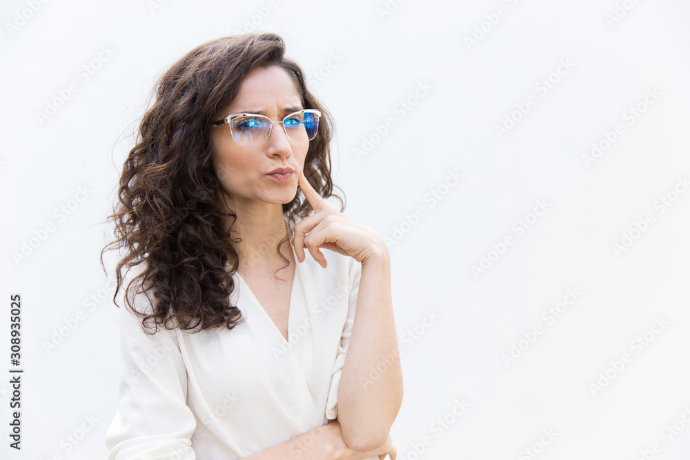 Strict frowning female professional in glasses touching face with finger, staring at camera. Wavy haired young woman in casual shirt standing isolated over white background. Teacher or mentor concept Strict frowning female professional in glasses touching face with finger, staring at camera. Wavy haired young woman in casual shirt standing isolated over white background. Teacher or mentor concept
