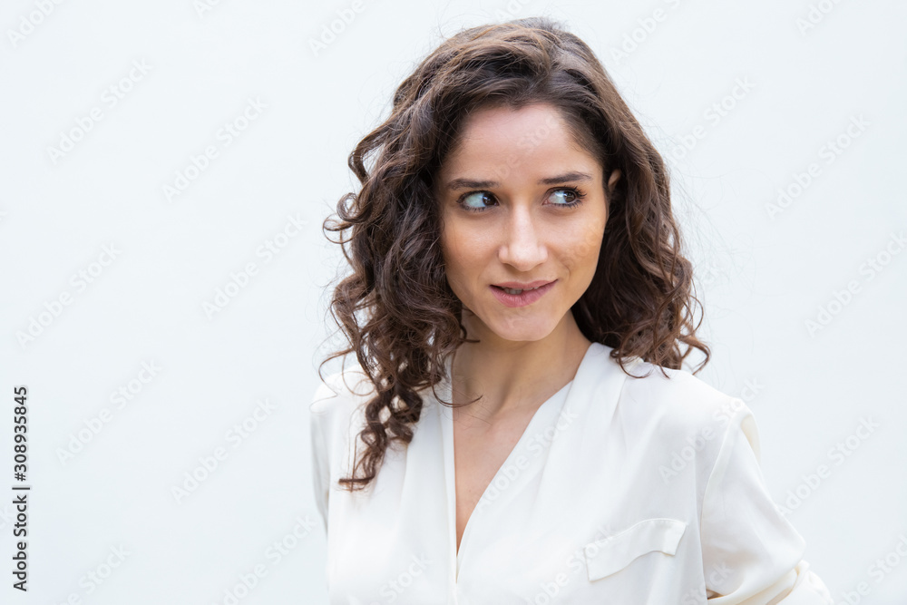 Positive flirty beautiful student girl biting lower lip, looking away at copy space. Wavy haired young woman in casual shirt standing isolated over white background. Female portrait concept Positive flirty beautiful student girl biting lower lip, looking away at copy space. Wavy haired young woman in casual shirt standing isolated over white background. Female portrait concept