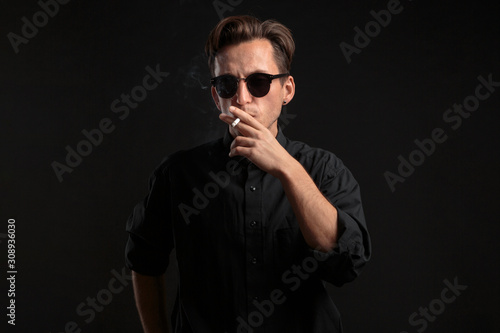 Stylish young man in a black t-shirt and sun glasses smoking a cigarette over black background.