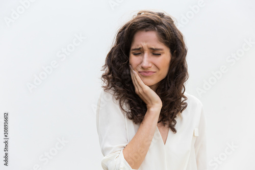 Frustrated unhappy woman suffering from toothache. Wavy haired young woman in casual shirt standing isolated over white background. Dental problem concept