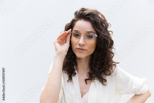 Serious positive female professional adjusting glasses. Wavy haired young woman in casual shirt standing isolated over white background. Corporate portrait concept