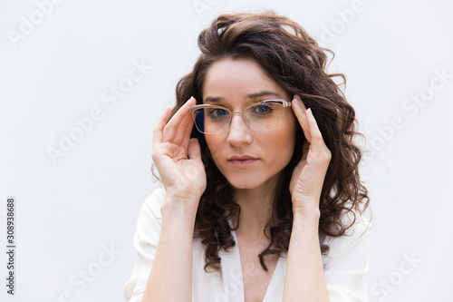 Portrait of serious positive female professional in glasses. Wavy haired young woman in casual shirt standing isolated over white background. Female portrait concept