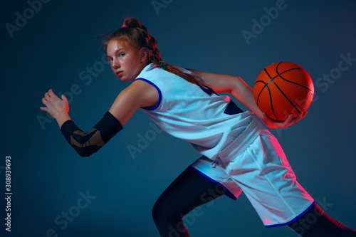 Flexibility. Young caucasian female basketball player on blue studio background in neon light, motion and action. Concept of sport, movement, energy and dynamic, healthy lifestyle. Training