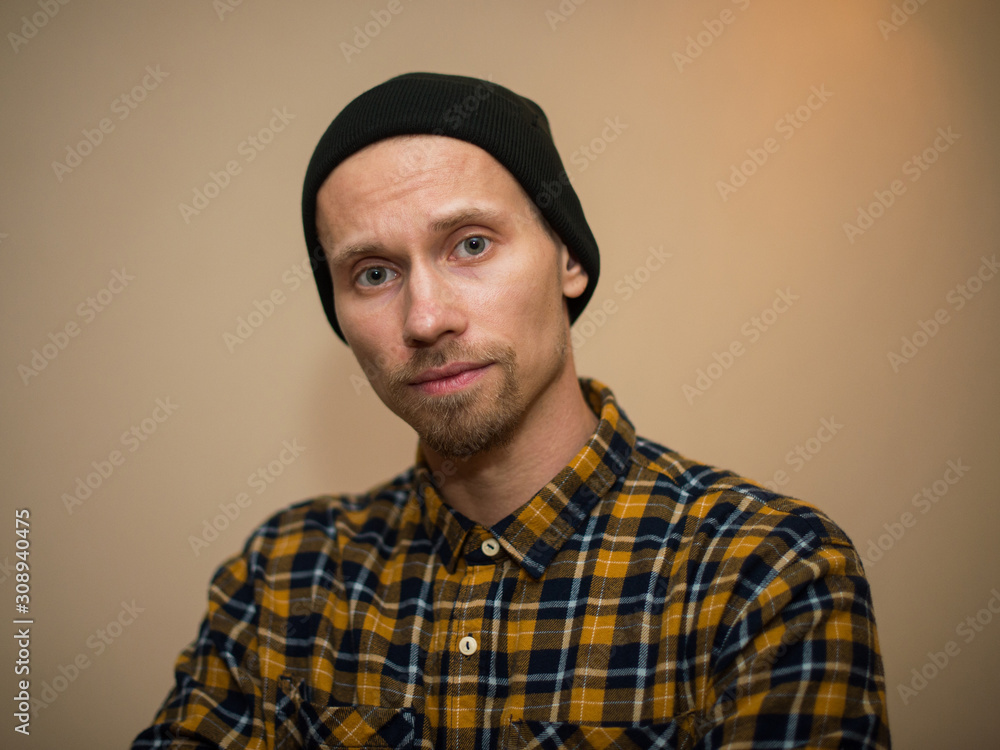 Young man, model of fashion, wearing a plaid shirt with a biege blind behind him. Guy in black hat with beard in casual style. Young man, model of fashion, wearing a plaid shirt with a biege blind behind him. Guy in black hat with beard in casual style.