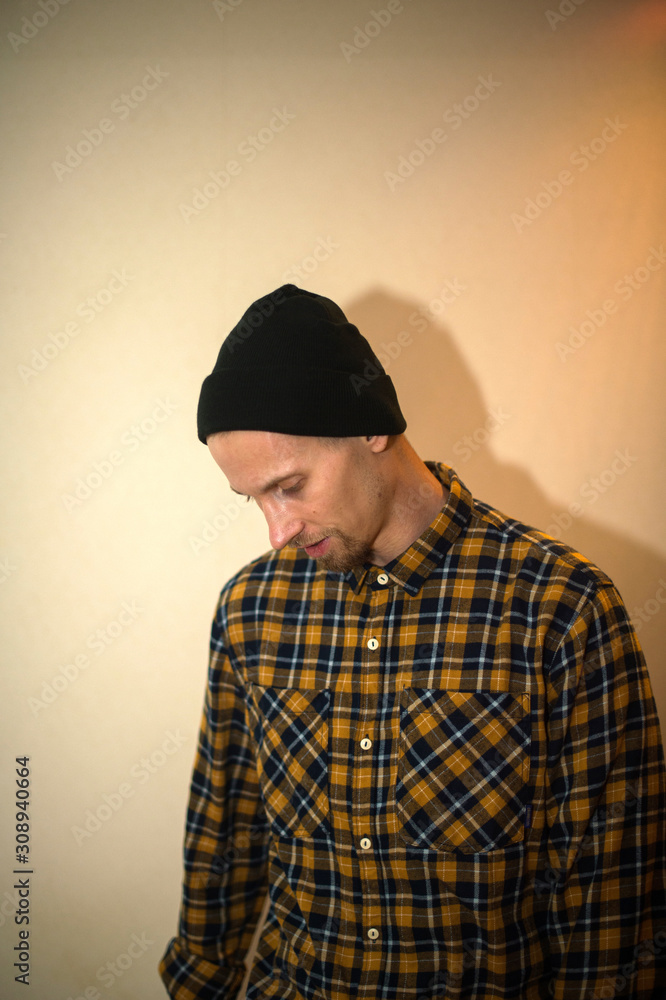 Young man, model of fashion, wearing a plaid shirt with a biege blind behind him. Guy in black hat with beard in casual style. Young man, model of fashion, wearing a plaid shirt with a biege blind behind him. Guy in black hat with beard in casual style.