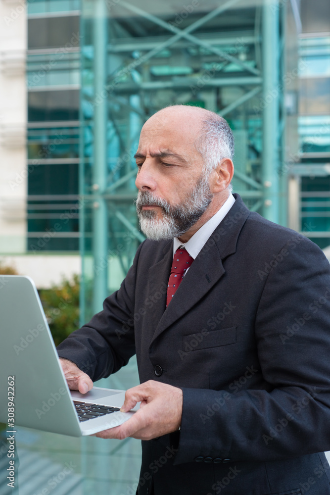 Focused mature businessman with laptop checking email near office building. Elderly man in formal suit and tie walking outside in city. Wireless technology concept Focused mature businessman with laptop checking email near office building. Elderly man in formal suit and tie walking outside in city. Wireless technology concept
