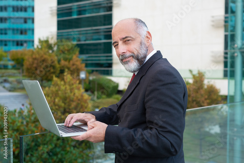Positive mature businessman using laptop near office building. Elderly man in formal suit and tie walking outside in city. Computer technology concept